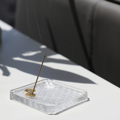 A brass moon-shaped incense holder with a plum blossom design on top, placed on a glass plate with a water wave pattern. The incense is lit, and smoke is rising from it.